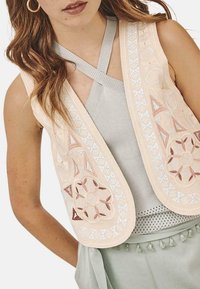 Chaleco bordado en beige con patrones florales y geométricos, con acentos plateados y una textura suave, superpuesto a una blusa de color gris claro.