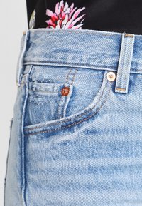 Vaaleansiniset korkeavyötäröiset farkut, joissa on näkyvät ompeleet, pieni etutasku ja kupariset niitit yksityiskohtina.