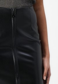 Fustă tip creion din piele artificială neagră, cu fermoar frontal, textură netedă și design ajustat. Prezintă o mână ținută lângă talie.