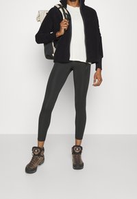 Czarna polarowa kurtka na zamek, biała koszulka, przylegające czarne legginsy i brązowe buty turystyczne z czarnymi akcentami. Plecak w ręce.