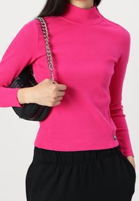 Přiléhavý rolák s dlouhým rukávem v jasně růžové rypsové tkanině, doplněný černou kabelkou s řetízkovým popruhem a texturovaným povrchem.