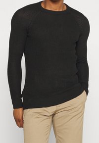 Svart stickad crewneck-tröja med ribbade ärmslut och nederkant, med en texturerad yta och figurnära silhuett. Kombinerad med beiga byxor.