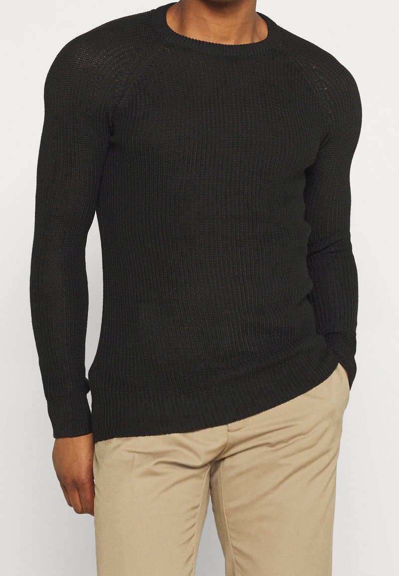 Svart stickad crewneck-tröja med ribbade ärmslut och nederkant, med en texturerad yta och figurnära silhuett. Kombinerad med beiga byxor.