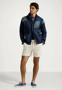 Denimjakke med varierende blå teksturer, knappelukking, kombinert med kremfargede shorts og blåmønstrede joggesko. Stripete skjorte under.