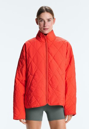 Žena oblečená v jasne oranžovej prešívanej bunde s vysokým golierom a olivovo zelených cyklistických šortkách, stojaca na svetlom pozadí.