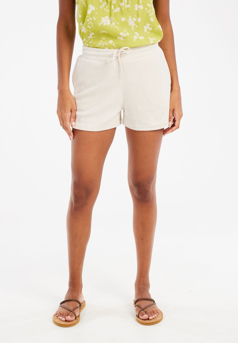 Shorts beige clair en tissu doux avec une taille élastique et un cordon de serrage, assortis à des sandales brunes à lanières.