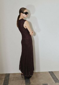 Abito in pizzo bordeaux con corpetto aderente e orlo svasato, abbinato a occhiali da sole neri oversize e scarpe con tacco alto su una superficie liscia.