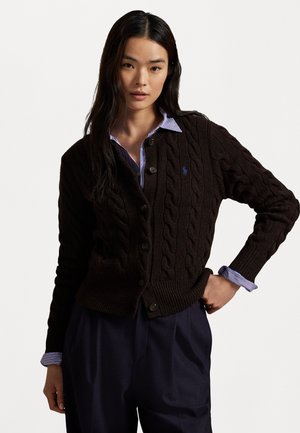 Brown cable-knit cardigan with a ribbed hem, featuring buttons down the front and a small logo. Underneath, a blue and white striped shirt.