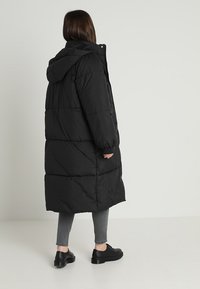 Abrigo tipo puffer negro con capucha, de diseño largo, textura acolchada y puños elásticos, combinado con pantalones grises y zapatos negros.