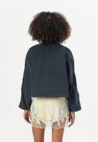 Cropped black denim jacket with wide sleeves. Back features a raw hem and top stitching details. Paired with a sheer, patterned skirt.