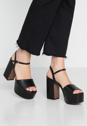 Czarne sandały na platformie z odkrytymi palcami, wykonane z materiału syntetycznego, z regulowanym paskiem wokół kostki i masywnym, szerokim obcasem. Widoczne postrzępione mankiety dżinsów.