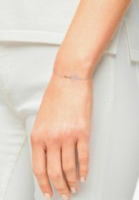 Bracciale in argento con chiusura a forma di cuore decorata con piccoli gemme trasparenti. Indossato su una mano con smalto naturale.