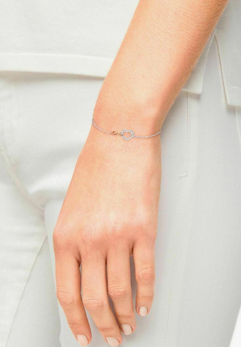 Bracciale in argento con chiusura a forma di cuore decorata con piccoli gemme trasparenti. Indossato su una mano con smalto naturale.