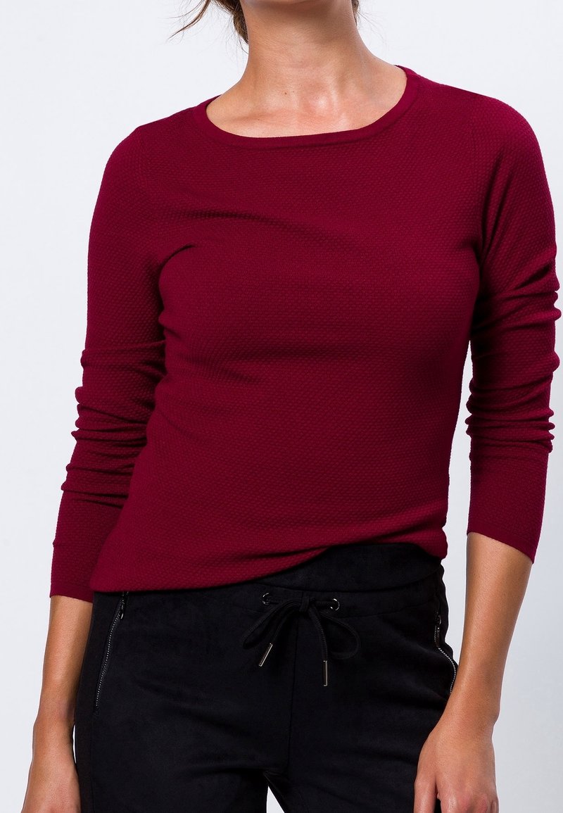 Chemise longue à manches longues texturée bordeaux avec un col rond, un design ajusté et des détails en côtelé. Associée à un pantalon noir à cordon de serrage.