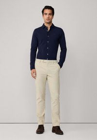 Man in een marineblauw overhemd met lange mouwen, beige broeken en donkerbruine schoenen, staand met één hand in de zak.