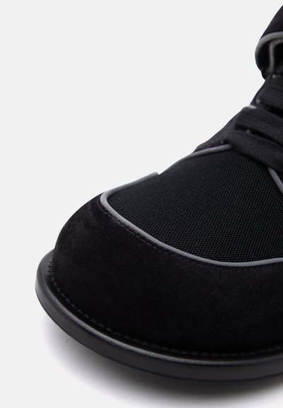 Κοντινό πλάνο στη μύτη ενός μαύρου αθλητικού παπουτσιού με πάνελς από σουέντ και δίχτυ, μαύρα κορδόνια και χοντρή λαστιχένια σόλα σε απλό φόντο.