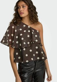 Jeune femme aux cheveux bouclés portant un chemisier marron à une épaule avec des pois blancs et un pantalon en cuir noir, regardant sur le côté.