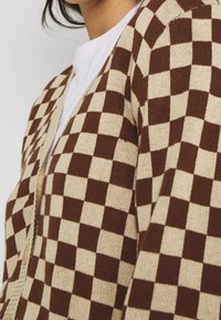 Braun- und cremefarbener karierter Strickcardigan mit V-Ausschnitt, gerippten Bündchen und offenem Design. Strukturierter Stoff erhöht den visuellen Kontrast.