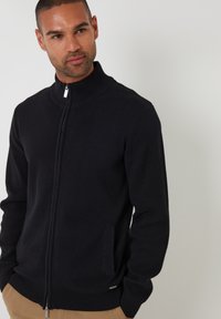 Maglione nero con zip e collo alto, con polsini a coste e una tasca, realizzato in materiale morbido. Logo discreto in fondo.