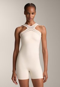 Beżowy bodysuit z teksturowanym, perforowanym wzorem krzyżujących się pasków. Gładki materiał, dopasowana sylwetka i krótkie nogawki.