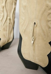 Tan suède cowboylaarzen met zwarte leren accenten, voorzien van gestikte patronen, een zijrits en een blokhak voor stabiliteit.
