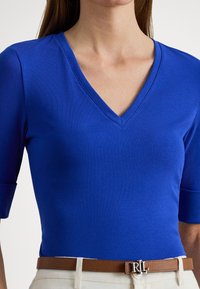 Žena oblečená v priliehavom kráľovsky modrom tričku s výstrihom do V a rukávmi dĺžky do lakťov, s hnedým koženým opaskom so striebornou prackou "RL".