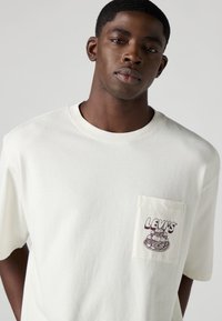 Λευκό βαμβακερό t-shirt με τσέπη στο στήθος που έχει ένα μοβ γραφικό σχέδιο στοίβαξης πιάτων. Λαιμόκοψη και κοντά μανίκια.