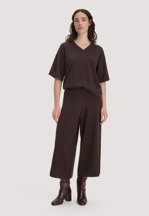 Frau mit dunklem welligem Haar, die ein lockeres, dunkelbraunes V-Ausschnitt-Kurzarmshirt, weit geschnittene knöchellange Hosen und dunkelbraune Stiefeletten trägt, stehend.