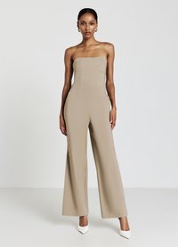 Combinaison sans bretelles beige avec un corsage ajusté, pantalon évasé et tissu texturé. Assortie à des chaussures à talons blancs.