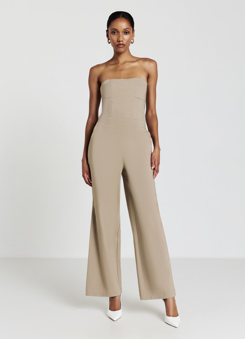 Combinaison sans bretelles beige avec un corsage ajusté, pantalon évasé et tissu texturé. Assortie à des chaussures à talons blancs.