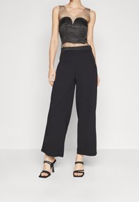 Czarny siatkowany top z dekoltem w kształcie serca i ozdobnym koralikiem, zestawiony z czarnymi spodniami o szerokich nogawkach i czarnymi szpilkami z paskami.