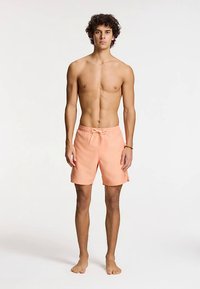 Broskvové plavkové šortky s elastickým pasem a šňůrkou. Materiál působí lehce. Model stojí bosý vzpřímeně.