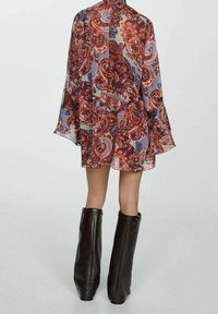 Blouse transparente et à motifs présentant des nuances de rouge et de bleu, avec des manches évasées. Associée à des bottes en cuir noir montantes jusqu'au genou.