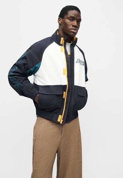 Νέος μαύρος άνδρας που φοράει μπλε σκούρο, λευκό και κίτρινο μπουφάν με φερμουάρ και τσέπες, σε συνδυασμό με μπεζ παντελόνι, ποζάροντας με τα χέρια στις τσέπες.