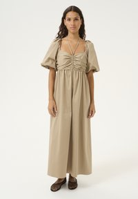 Robe maxi beige avec des manches bouffantes et un buste froncé. Fabriquée en tissu lisse, avec un décolleté noué et une coupe décontractée. Chaussures en mesh noires.