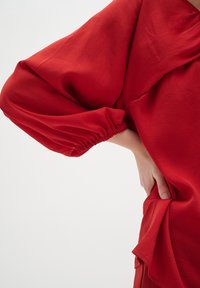 Czerwona bluzka z bufiastymi rękawami i elastycznymi mankietami, charakteryzująca się luźnym krojem i gładką fakturą, częściowo pokazana z ręką na odzieży.