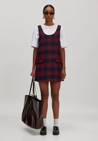 Marineblaues und rotes kariertes Kleid mit Rundhalsausschnitt und zwei vorderen Taschen; über einem weißen T-Shirt getragen. Das Modell trägt eine glänzende burgunderrote Tote-Tasche.