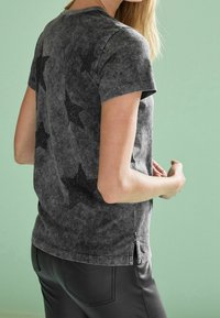 Sort acid-wash t-shirt med korte ærmer, der har glitrende sorte stjerne mønstre på ryggen og sideslids ved kanten.