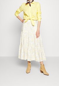 Keltainen pellavainen napillinen paita, joka on sidottu vyötärölle, yhdistetty valkoiseen kukkakuosisessa kerroksellisessa hameeseen ja vaaleisiin nilkkasaappaisiin. Huivikoriste kaulassa.