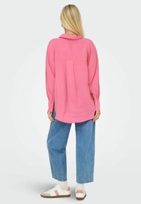 Camicia rosa a maniche lunghe con colletto, caratterizzata da una piega sul retro. Abbinata a jeans larghi di un blu chiaro e sneakers bianche.