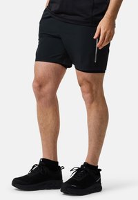 Mustat urheilushortsit, joissa on sileä ja kevyt kangas, sivutasku vetoketjulla. Yhdistetty mustiin, tekstuurisiin urheilukenkiin.