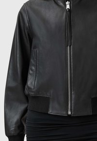 Chaqueta bomber de cuero negro con cremallera frontal plateada, puños acanalados y bolsillos laterales. Textura suave con un diseño ajustado.