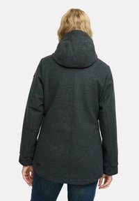 Blondi henkilö, joka on pukeutunut tummanharmaaseen huppariin ja sinisiin farkkuihin, kuvattuna selkäpuolelta yksinkertaista valkoista taustaa vasten.