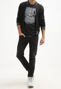 Černý zipový svetr přes černé tričko s potiskem, které obsahuje bílé linie a text, v kombinaci s černými kalhotami a bílými teniskami.