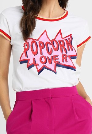 Vrouw draagt een wit T-shirt met rode en gele bies, met rode en roze tekst 'Popcorn Lover', gecombineerd met felroze broek met hoge taille.