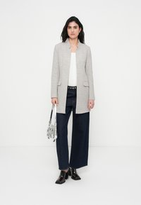 Μακριά γκρι ανοιχτόχρωμη υφασμάτινη ζακέτα χωρίς γιακά, με δύο εμπρόσθιες τσέπες, συνδυασμένη με σκούρα τζιν φαρδιάς γραμμής και γυαλιστερά μαύρα μποτάκια μέχρι τον αστράγαλο.