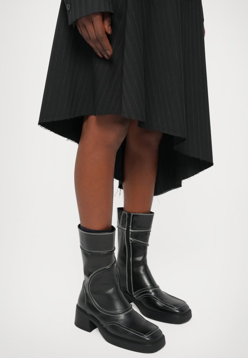 Schwarze Lederstiefeletten mit quadratischer Zehenpartie, Blockabsatz, weißer Naht und umgeschlagenem Design, kombiniert mit einem gestreiften schwarzen Rock.