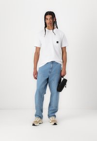 Tricou alb din bumbac cu buzunar frontal, pantaloni din denim de un albastru deschis cu cusături vizibile și adidași bej. Pungă neagră ținută în mâna stângă.
