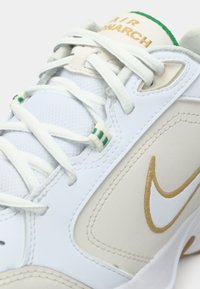 Witte sportschoen met beige accenten, bovenwerk van gestructureerd leer en mesh, voorzien van een groene logosticker, witte veters en een gouden geborduurd label.