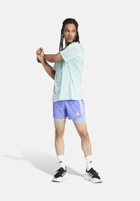 Persona che si allunga le braccia indossando una maglietta sportiva azzurro chiaro, pantaloni viola, calze bianche Adidas e scarpe nere Adidas su uno sfondo bianco.